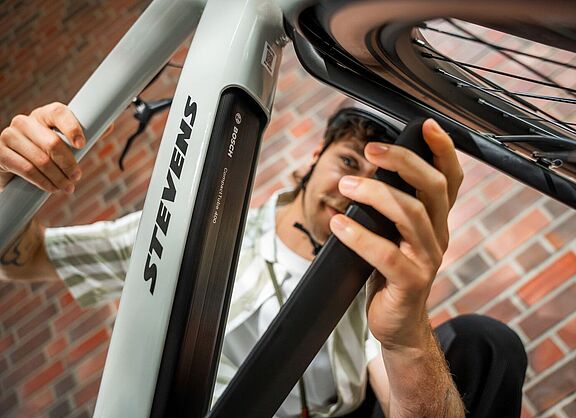 Ein Mann öffnet die Akku-Abdeckung an einem grauen Elektrorad, von schräg unten aufgenommen. 